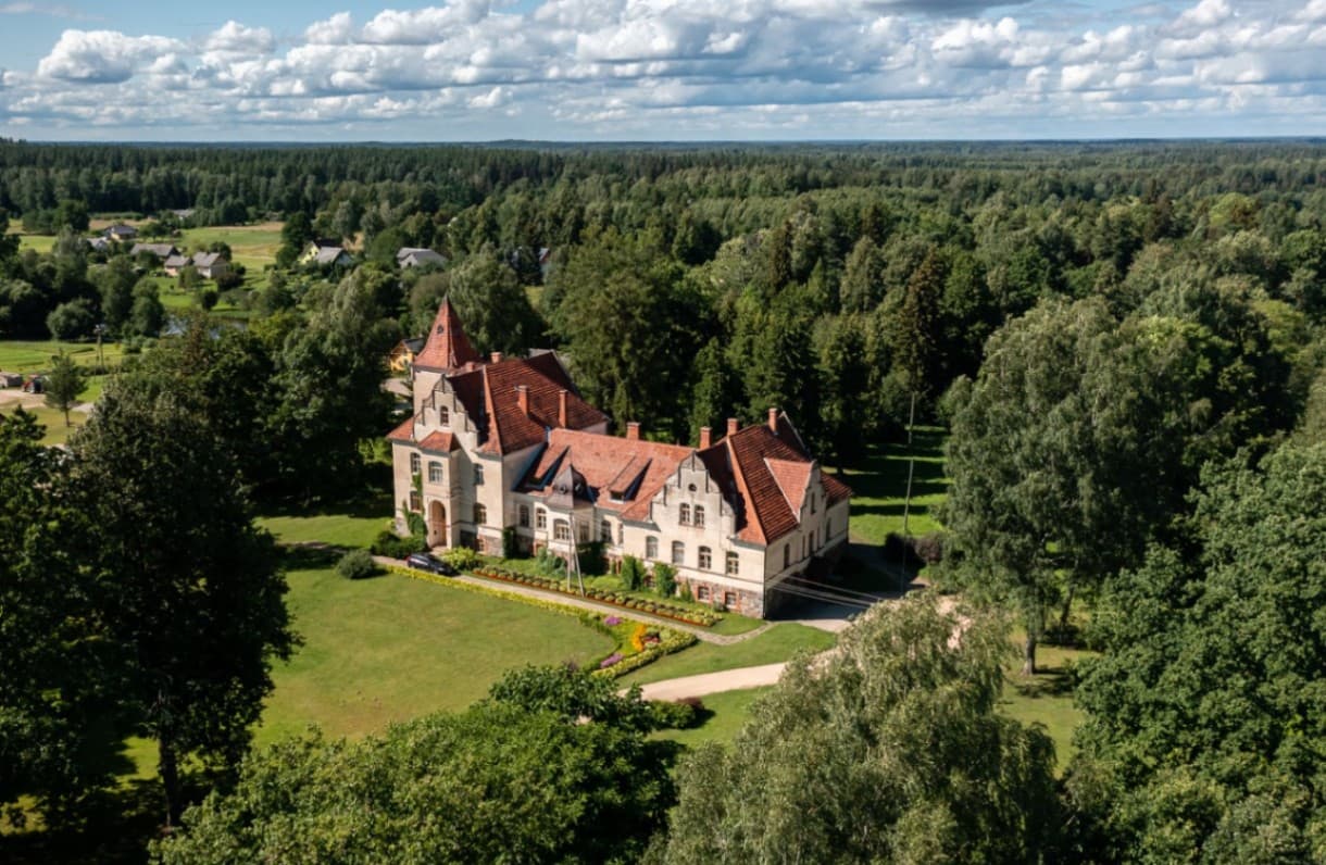A bird’s-eye view photograph with a historic manor building in the centre of the image. The building has a light plaster facade, a stone plinth, a red tiled roof and a tower section. It is su