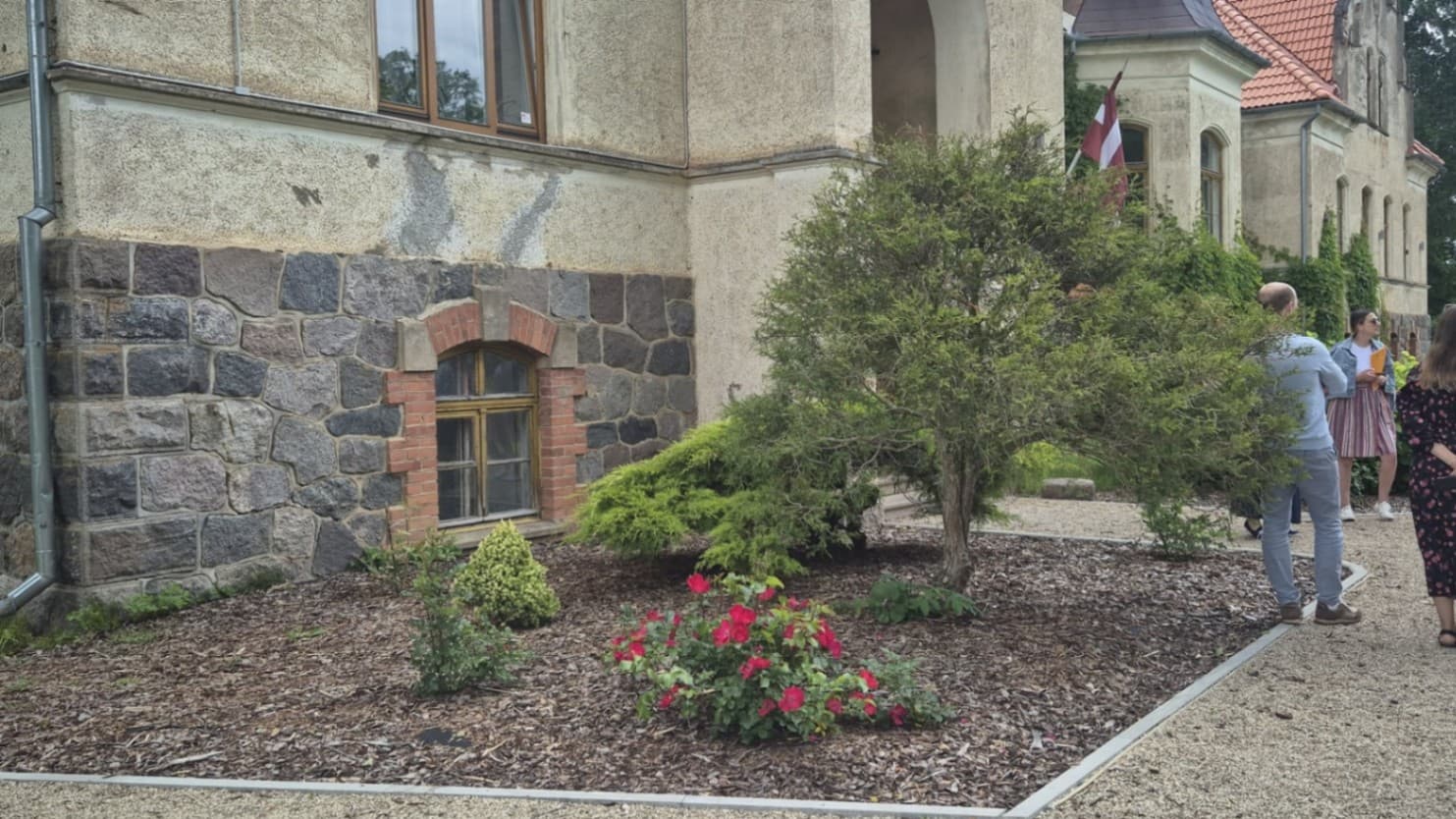 The photograph was taken from the corner of the manor building. In the centre of the image there are plantings next to the manor building with a mulched flower bed, conifers, ornamental shrub