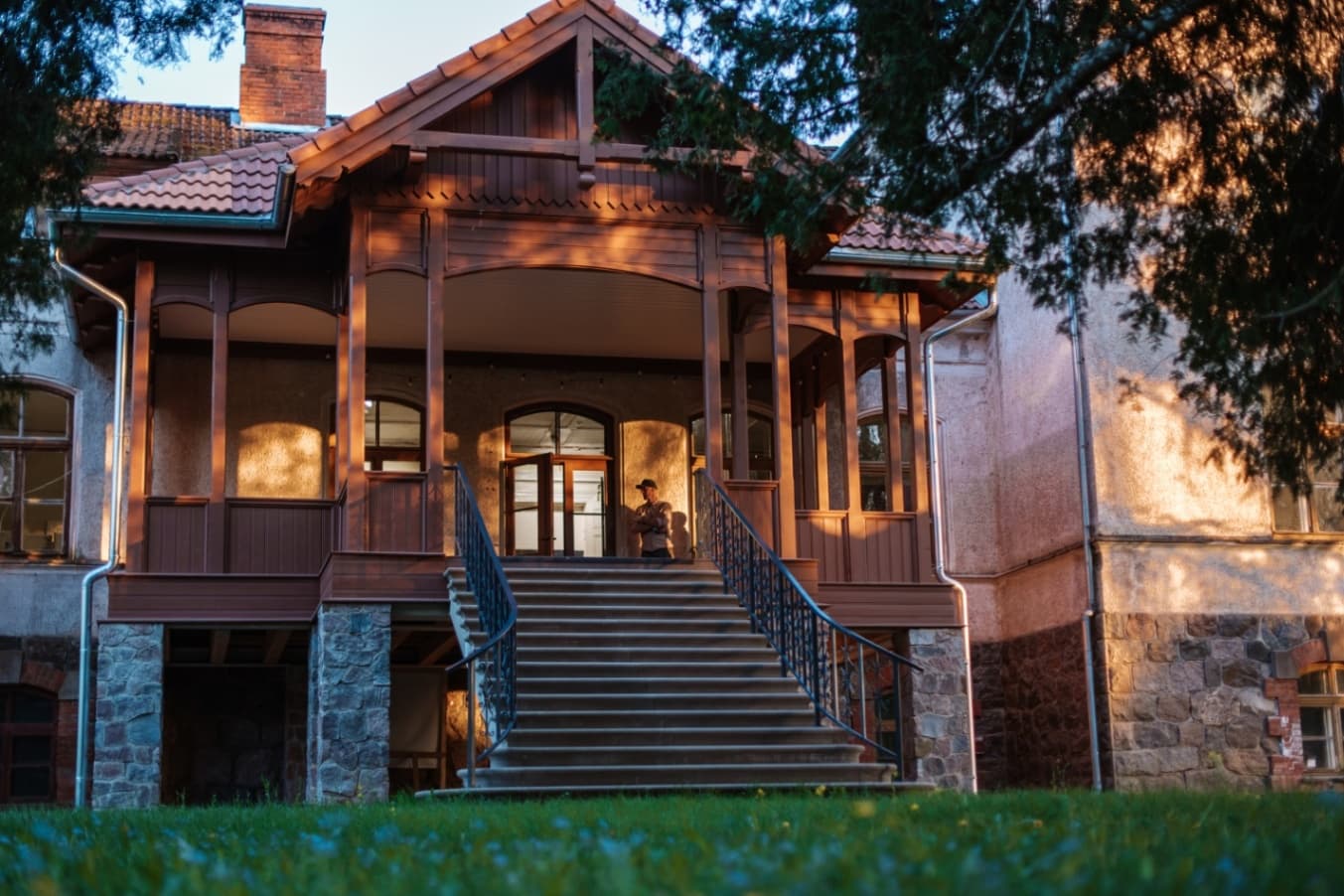 The entrance area of the manor building with wide steps, metal railings, a wooden veranda with a red tiled roof. A person is standing by the door, while the facade is lit by warm evening ligh