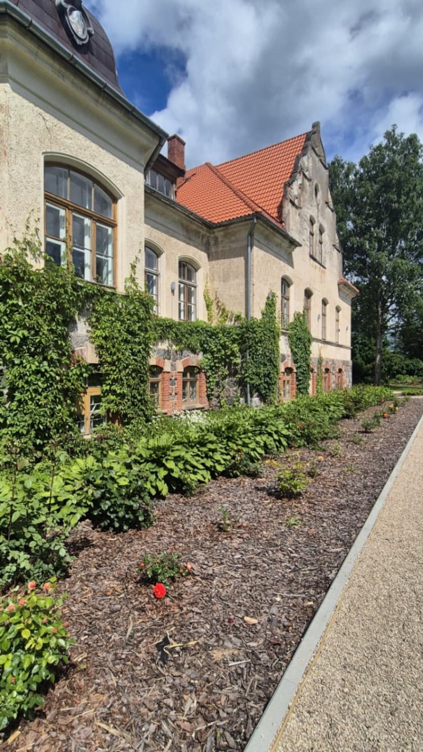 The side facade of the manor building has light plaster, a red tiled roof, wooden windows and a stone plinth with brick edging. Along the building there is a mulched flower bed with climbing 