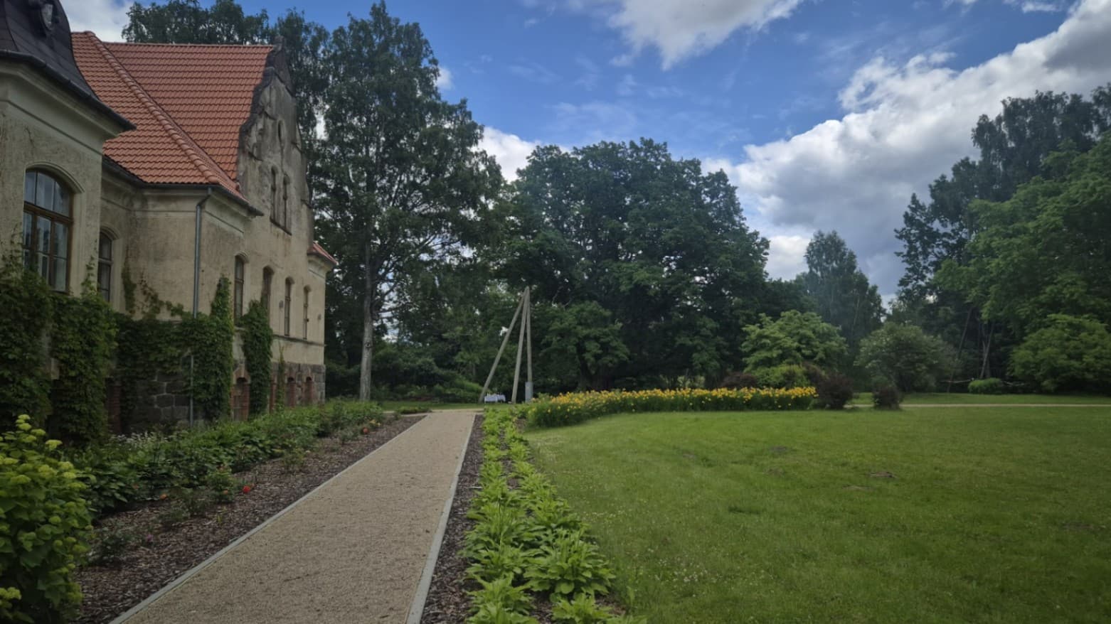 A light gravel path runs along the manor building and the well-kept park. On the left, the facade of the historic building with a stone plinth, climbing plants and flower beds is visible, whi
