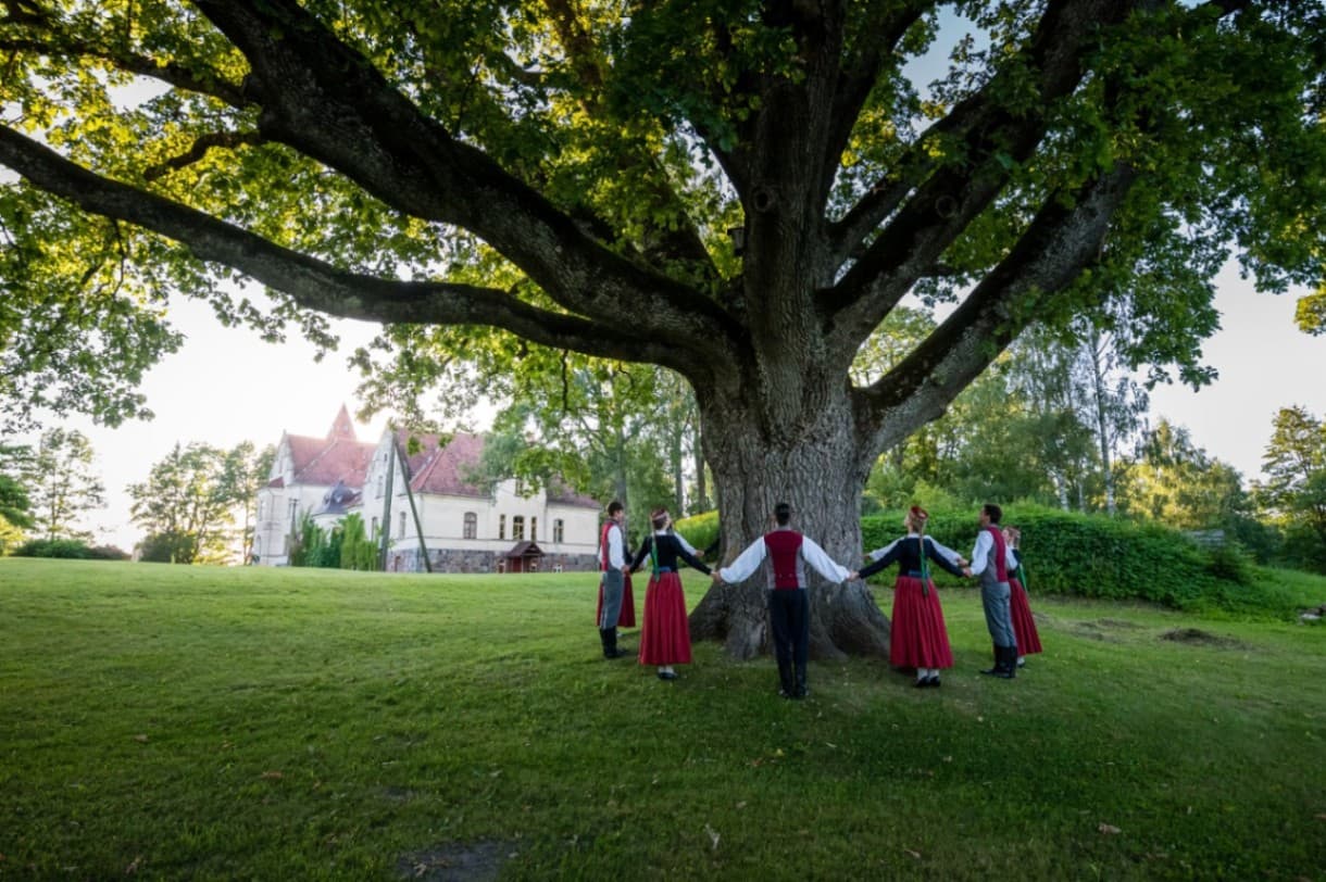 A large, spreading oak tree in the manor park, around which people in folk costumes are standing hand in hand. In the background there is a historic manor building with a light façade and a r