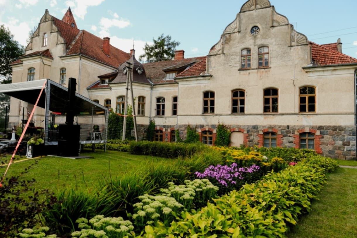 In front of the manor building there is an event stage with sound and lighting equipment. In the foreground there are well-kept decorative flower beds with hostas, ornamental grasses, hydrang